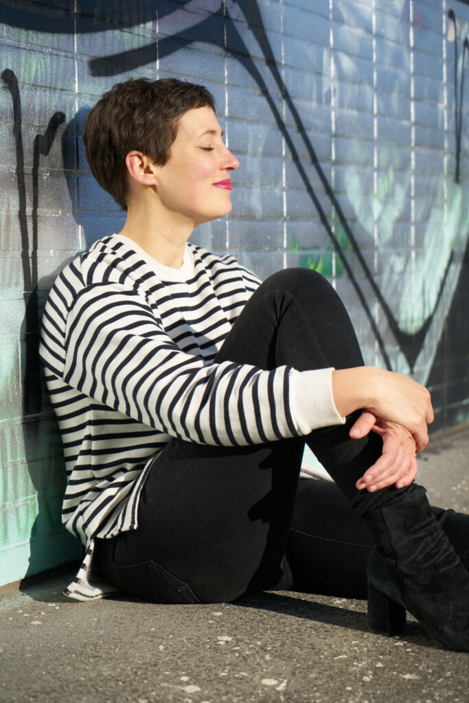 Frau mit gestreiftem Pullover sitzt am Boden mit geschlossenen Augen - Beitragsbild für das Interview mit Nuria Fischer